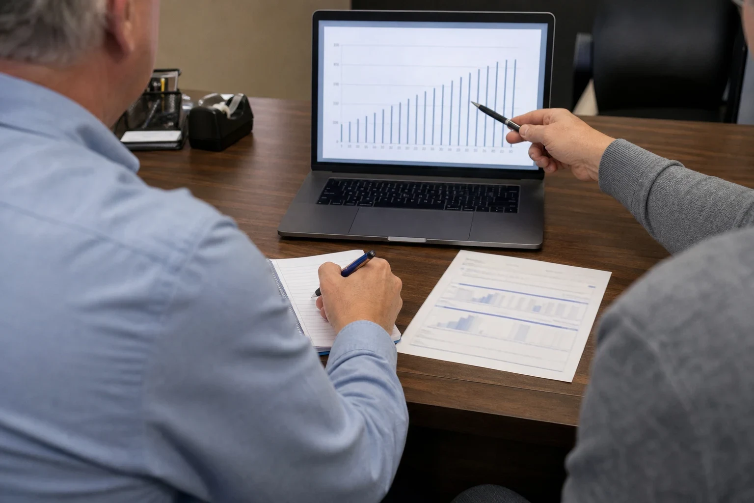 Over-the-shoulder view of an agent pointing a pen at a rising bar chart on a laptop while a gray-haired client takes notes beside a printed chart at an office desk