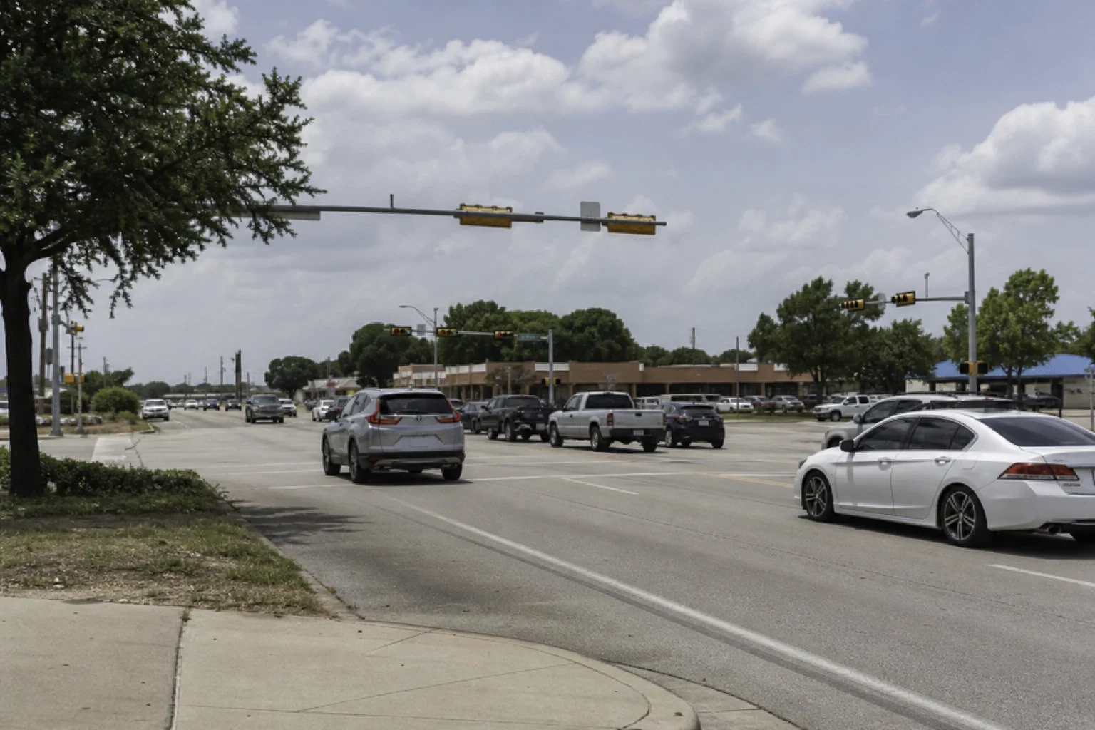 Wide suburban intersection with traffic signals, cars and pickup trucks, a brick strip shopping center and live oak trees under a partly cloudy sky