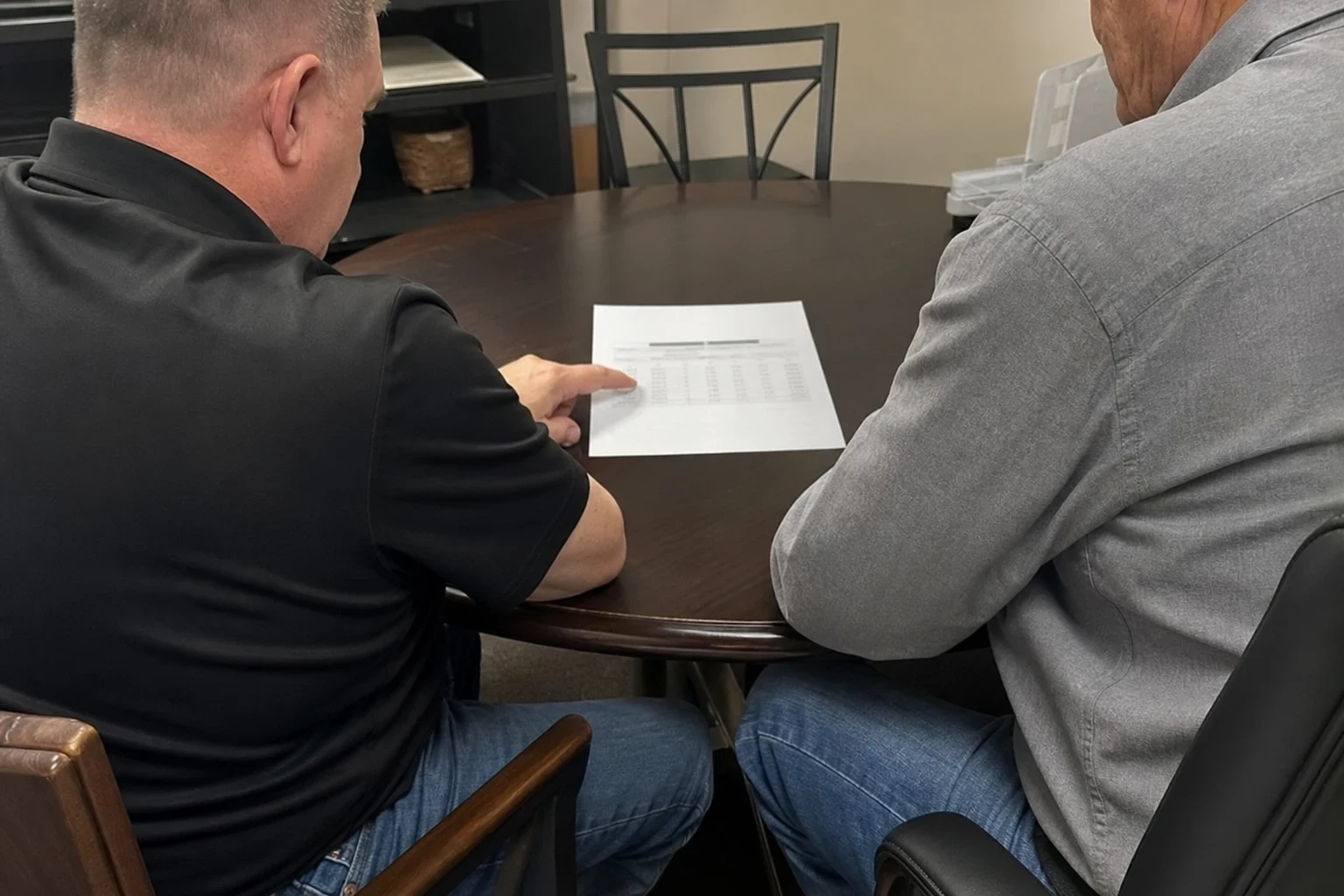 Two men seated at a small round wooden table in an office, one pointing at a printed page of figures as they review it together.