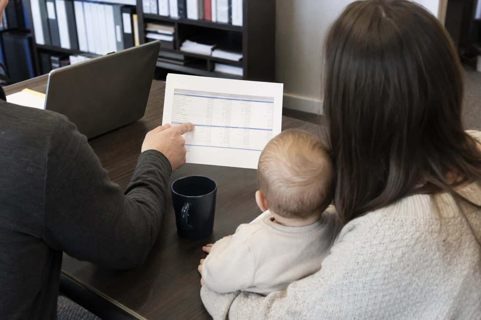 Agent at an office desk pointing to a printed comparison sheet for a woman holding a baby, with a laptop, dark coffee mug and shelves of binders behind